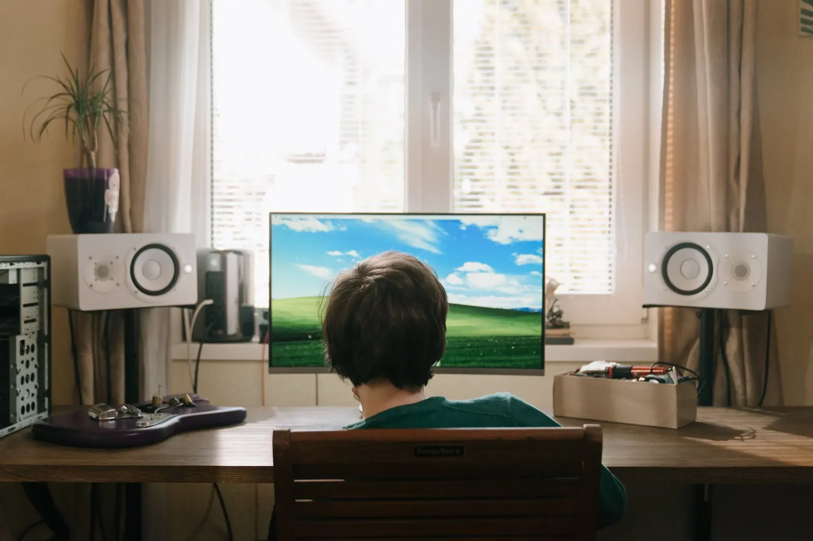 The image shows a person seated at a computer desk with a large flat-screen TV mounted on the wall behind them. The room has a homely feel, with a window allowing natural light to enter, and various objects such as a potted plant, speakers, and additional items on the desk are visible.